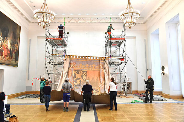 Vom Landtag ins Museum: Der Wandteppich aus dem Bayerischen Landtag wird im Museum des Hauses der Bayerischen Geschichte in Regensburg ausgestellt werden. Im Juli 2017 wurde der rund sieben mal sieben Meter große Gobelin fachmännisch abgenommen. Der Bildteppich hing von 1950 bis 2004 im alten Plenarsaal, nach dessen Umbau zog er in den Senatssaal um. 