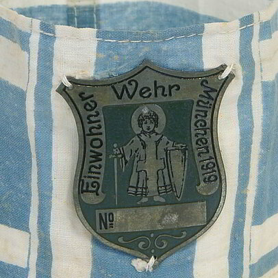 Ab Frühjahr 1919 entstanden in Bayern zahlreiche sog. Einwohnerwehren, gefördert v.a. vom späteren Ministerpräsidenten Gustav Ritter von Kahr. Die Einwohnerwehren waren ein dauernder Streitpunkt zischen bayerischer Landesregierung, Reichsregierung und den Siegermächten. Im Juni 1921 wurden sie auf Druck der Entente verboten.
In der Sammlung befindet sich diese weiß-blaue Armbinde der Münchner Einwohnerwehr. Aufgenäht eine Art Erkennungsmarke in Form eines Schildes aus Metall, darauf abgebildet das Münchner Kindl mit Schwert und Schild, Umschrift „Einwohner Wehr München 1919“.