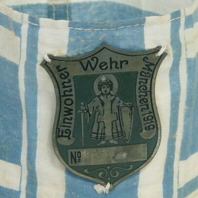 Ab Frühjahr 1919 entstanden in Bayern zahlreiche sog. Einwohnerwehren, gefördert v.a. vom späteren Ministerpräsidenten Gustav Ritter von Kahr. Die Einwohnerwehren waren ein dauernder Streitpunkt zischen bayerischer Landesregierung, Reichsregierung und den Siegermächten. Im Juni 1921 wurden sie auf Druck der Entente verboten.
In der Sammlung befindet sich diese weiß-blaue Armbinde der Münchner Einwohnerwehr. Aufgenäht eine Art Erkennungsmarke in Form eines Schildes aus Metall, darauf abgebildet das Münchner Kindl mit Schwert und Schild, Umschrift „Einwohner Wehr München 1919“.